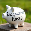 Das Bild zeigt ein weißes Sparschwein aus Keramik mit der Aufschrift „Nichtraucher Kasse“ in schwarzer Schrift. Das Sparschwein hat eine klassische Form mit spitzen Ohren und einem Schlitz oben, um Geld einzuwerfen. Es steht auf einem Stein oder einer festen Unterlage im Freien, wobei im Hintergrund eine grüne, verschwommene Wiesenlandschaft zu sehen ist. Das Bild vermittelt einen frischen, positiven Eindruck, der möglicherweise mit Nichtrauchen und Sparen assoziiert werden soll.