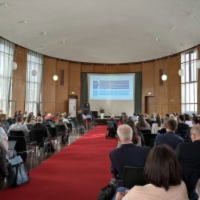 Das Bild zeigt eine gut besuchte Veranstaltung in einem großen, hellen Saal mit Holzverkleidung und hohen Fenstern. Die Teilnehmenden sitzen in Reihen und schauen auf eine Bühne mit rotem Teppich, auf der eine Person einen Vortrag hält. Hinter der Person wird eine Präsentation auf eine große Leinwand projiziert.